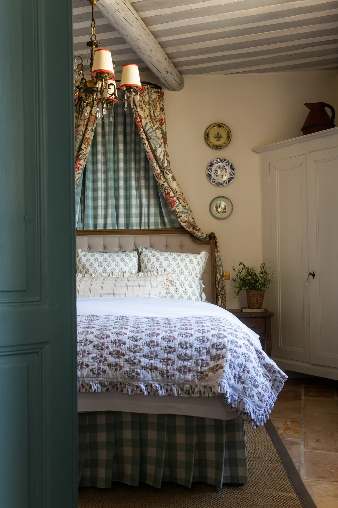 Chambre Two — a queen bed under a green gingham canopy with floral chintz drapes, a white armoire alongside, and terracotta tiles beneath a whitewashed beamed ceiling