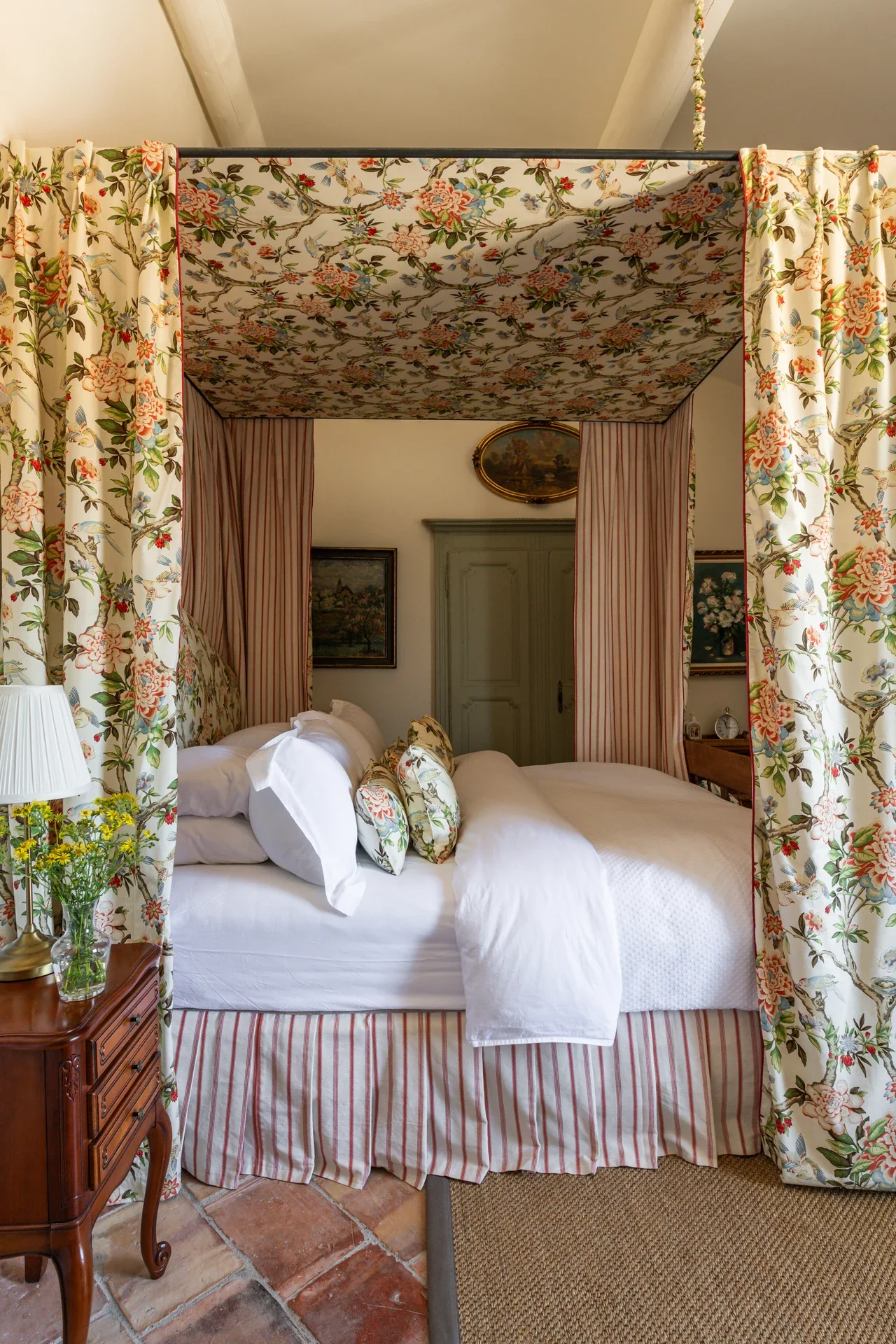 Chambre Three — a four-poster bed canopied in floral chintz, its pink ticking-stripe curtains drawn back over white linen and a matching bed skirt, on terracotta tiles