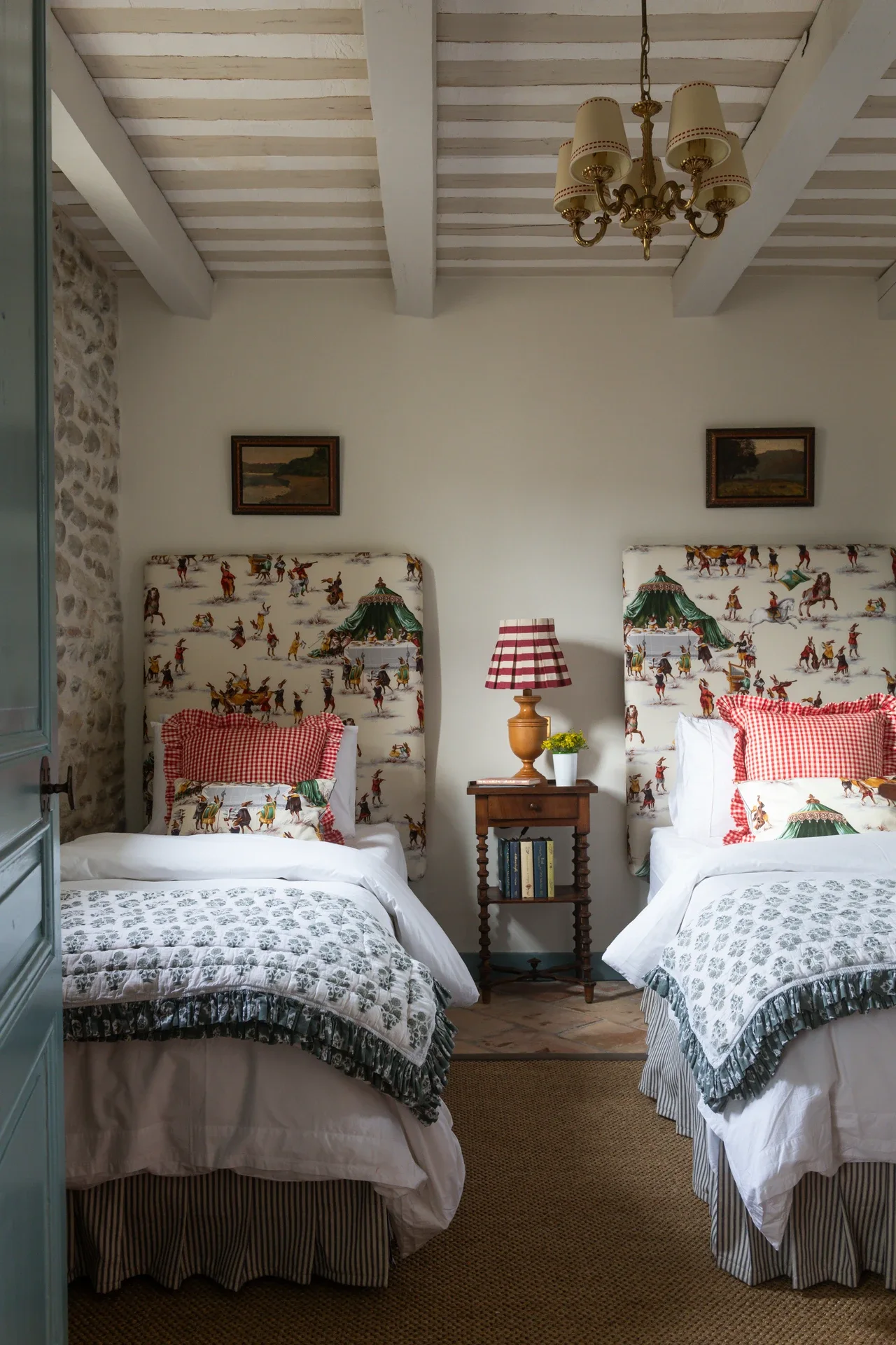 Chambre Four — two extra-long single beds with hunt-scene headboards on either side of a walnut bedside table, red gingham cushions on white linen, beneath a whitewashed beamed ceiling